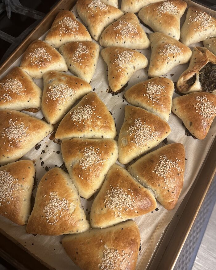 A sheet pan of freshly baked spinach fatayer — golden three-cornered hand pies, each brushed with egg wash and scattered with sesame and nigella seeds.