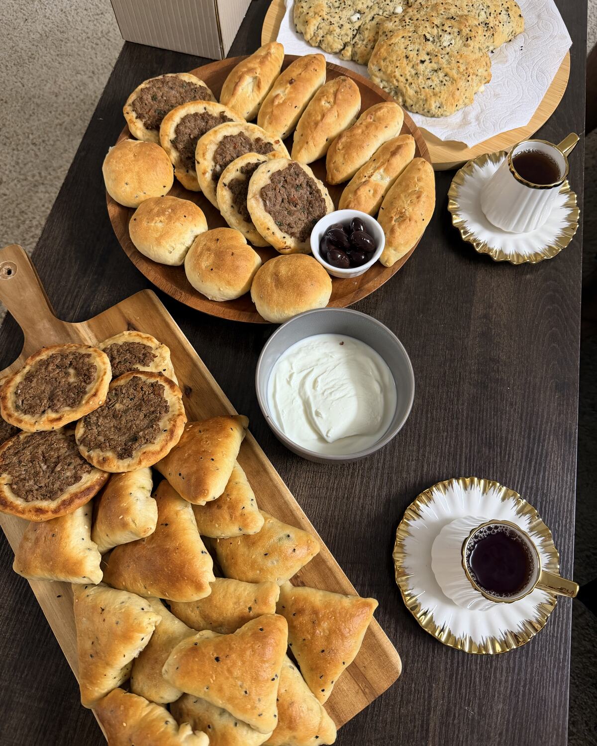 An overhead table spread of Palestinian baked goods: a wooden platter of meat sfiha and sesame-topped rolls beside a board of spinach fatayer triangles, a bowl of labneh, black olives, a sesame and nigella flatbread, and two gold-rimmed glasses of Arabic tea.