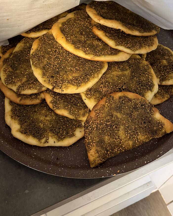 A stoneware platter piled with za'atar man'oushe — round flatbreads baked with a thick, seeded green za'atar and olive oil topping, edges puffed and golden.